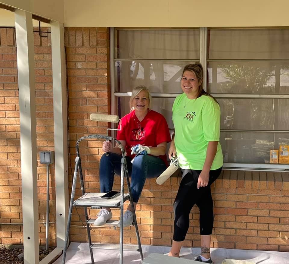Two women on a porch, one seated with a walker, one standing beside her in a bright green shirt.