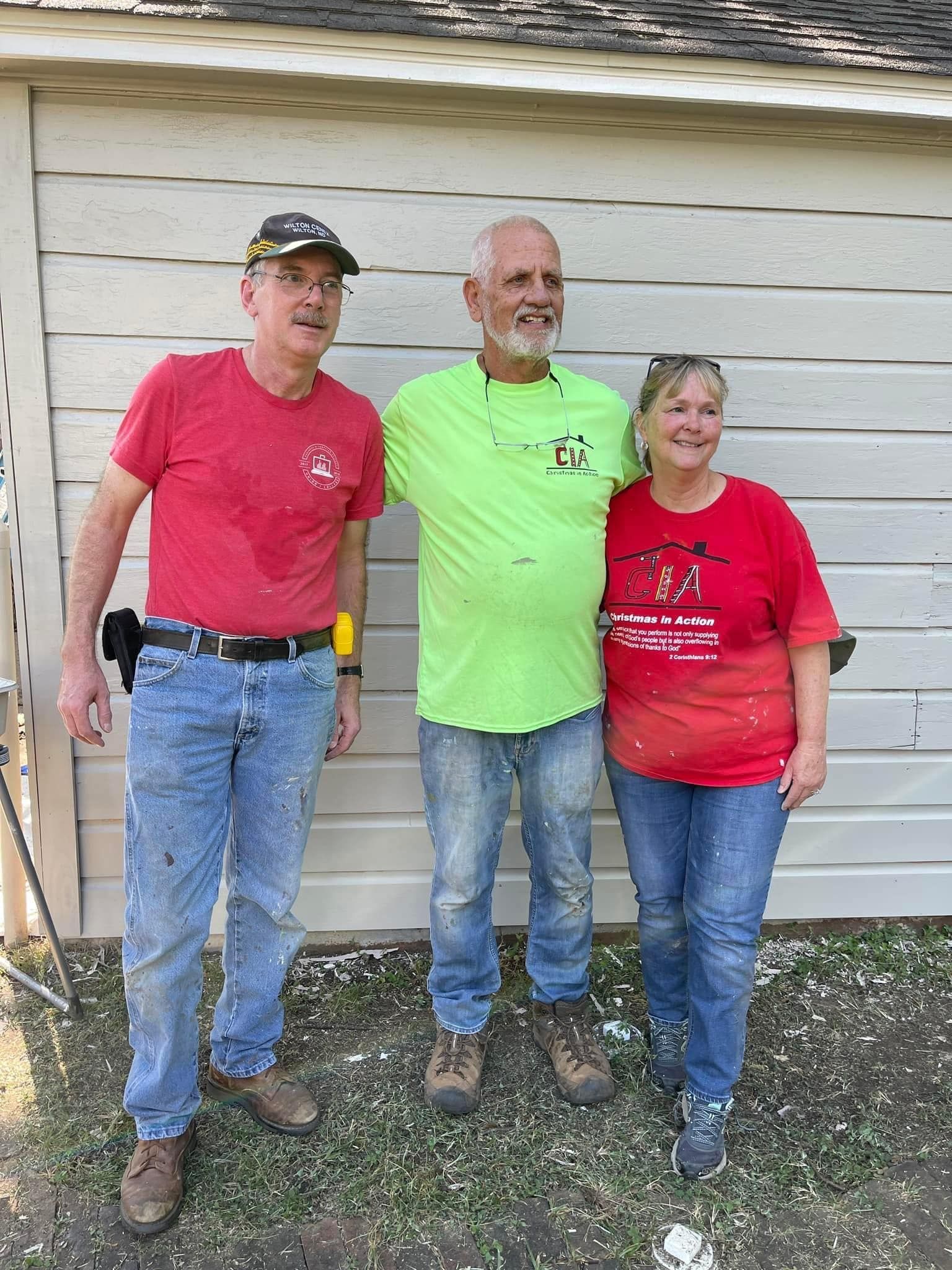 Three adults standing outdoors against a white wooden wall, wearing red and neon green shirts and jeans.