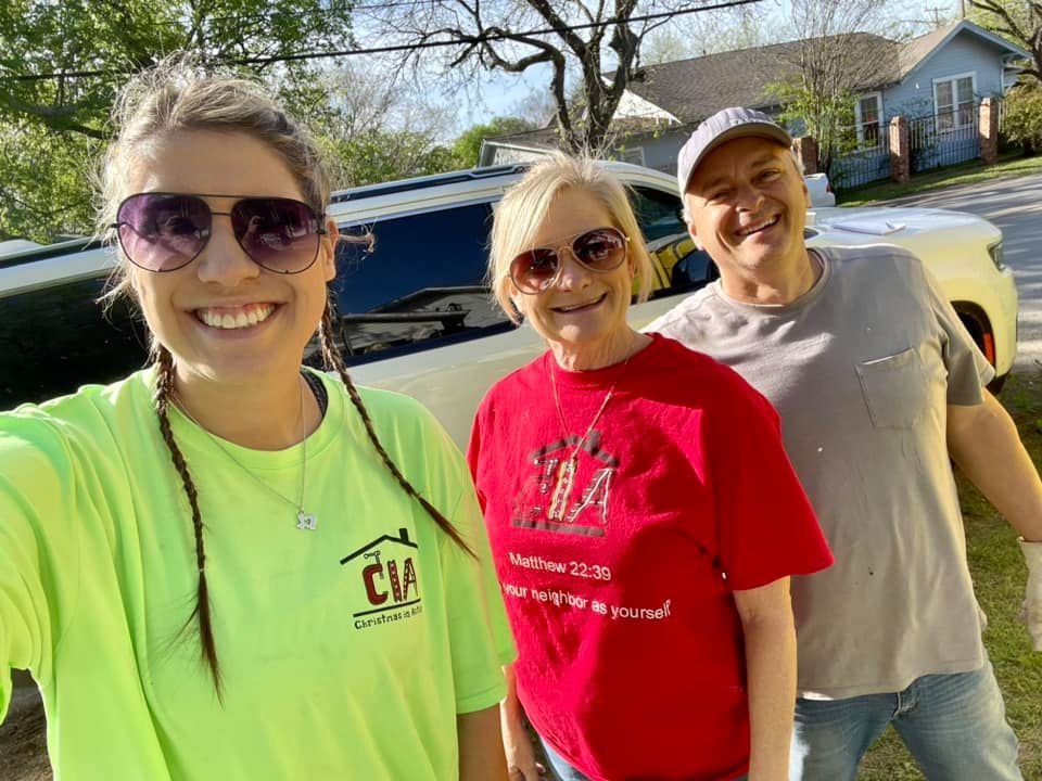 Three smiling people standing outdoors in sunlight, posing in front of a house and truck.