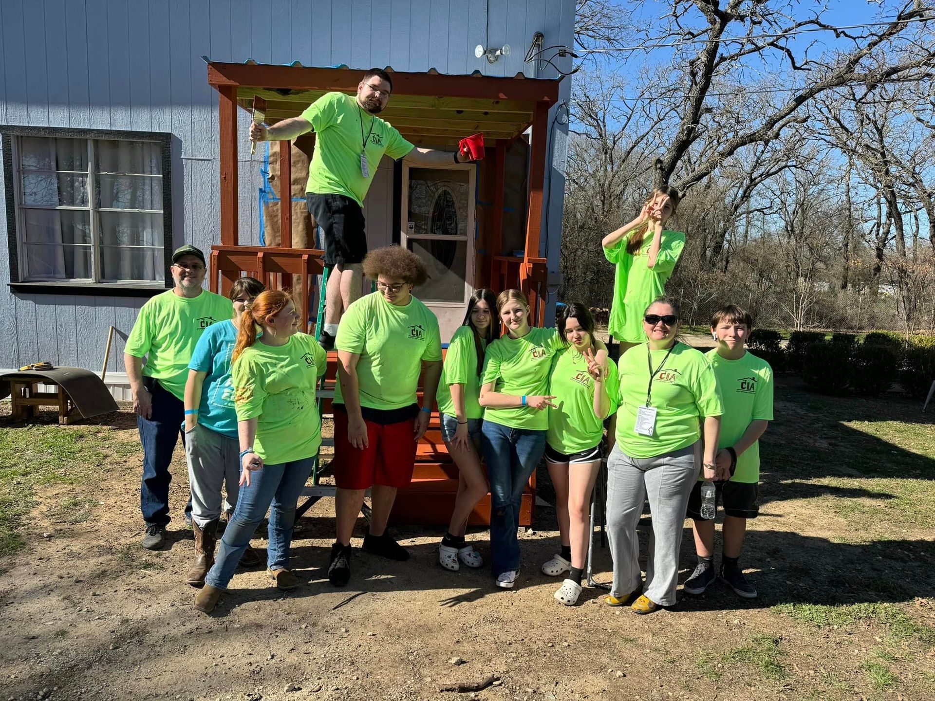 Group in neon green shirts posing on a porch in front of a blue house, with one person jumping nearby