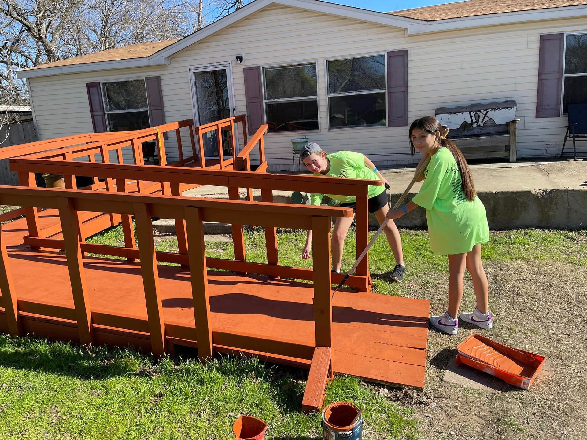 People working on a large orange wooden ramp outside a white house, with tools on the grass