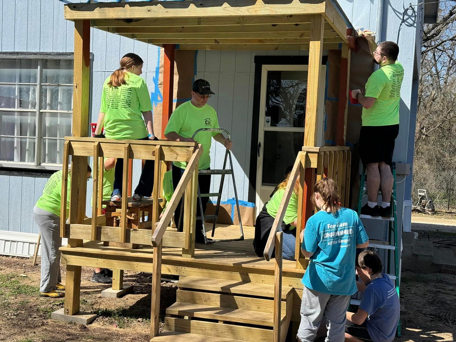 Volunteers in neon green shirts building a porch on a house, with a man on the roof and others on ladders.