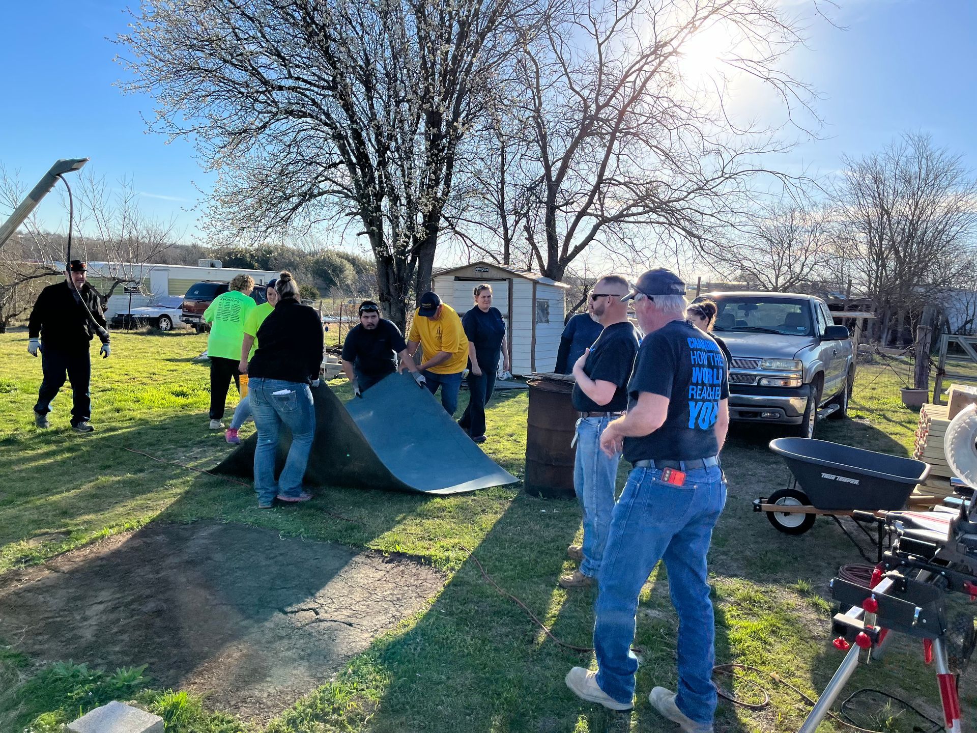 People setting up a tent in a sunny park, with cars and bicycles nearby.