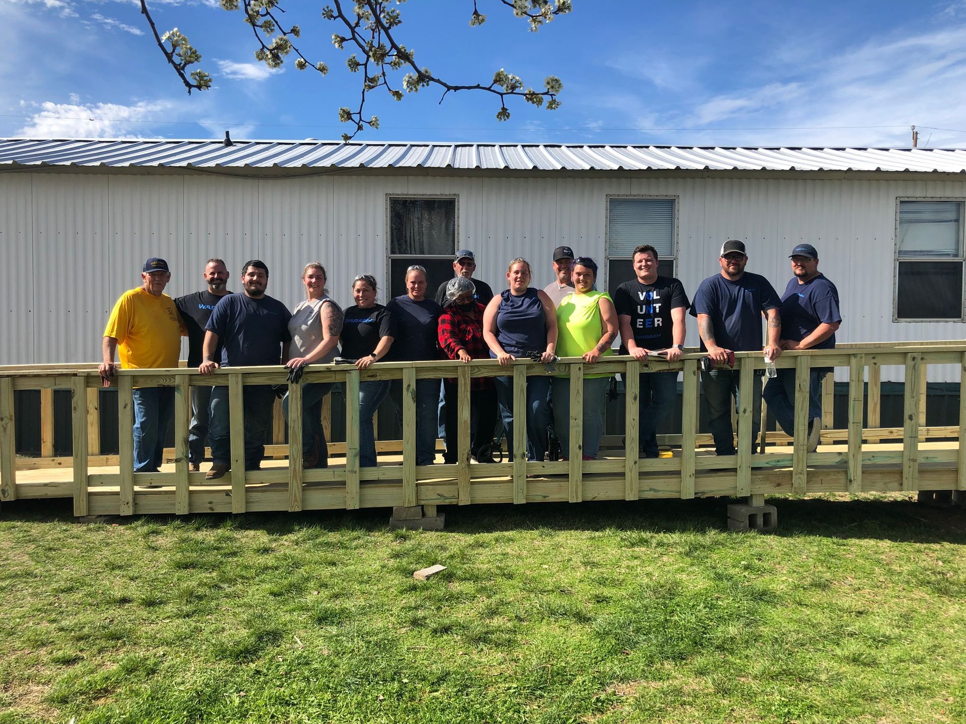 Group of people standing on a wooden deck in front of a white building on a grassy lawn.