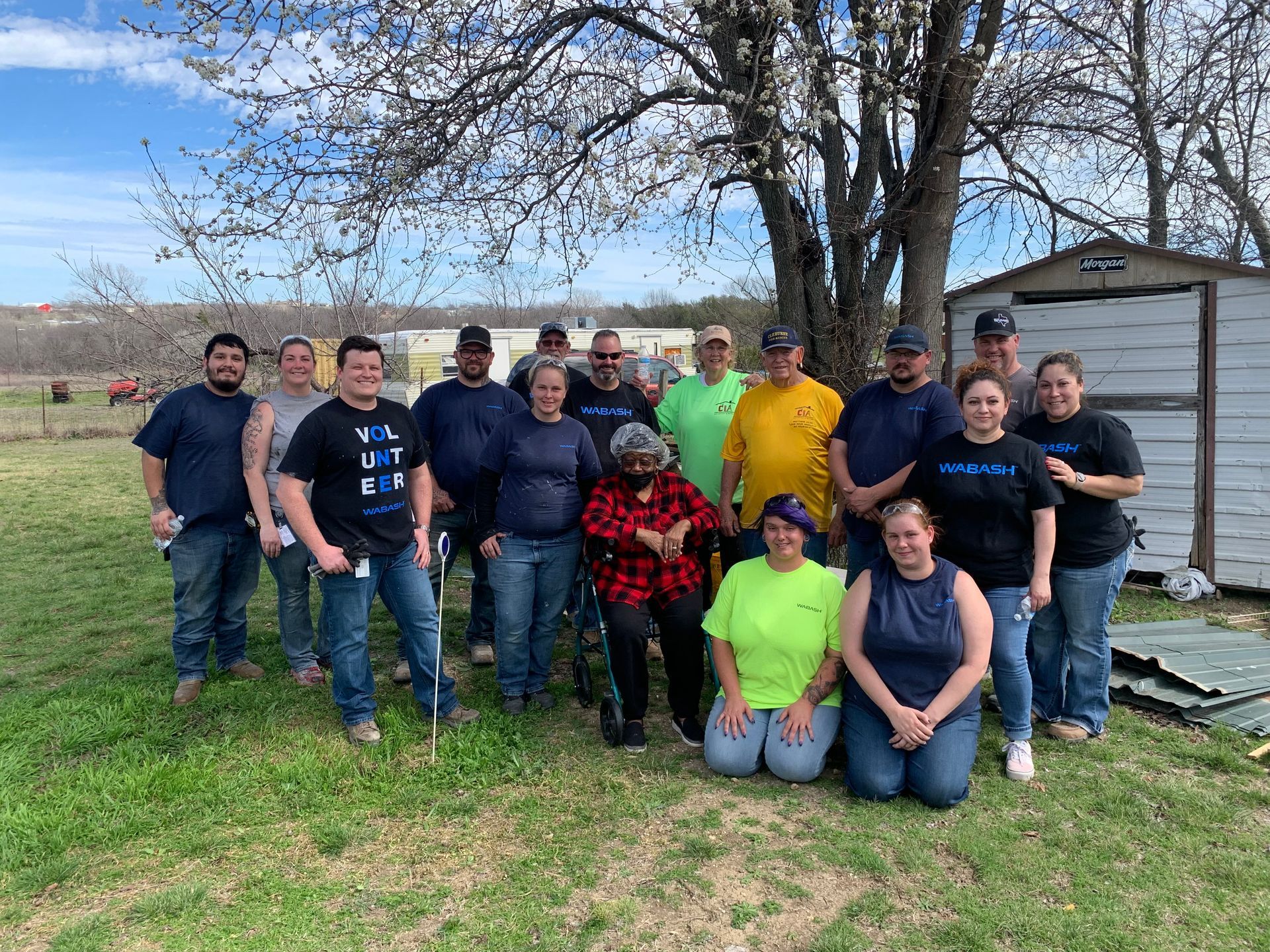 Group of people posing outdoors on a grassy lawn beside a small shed and leafless tree.