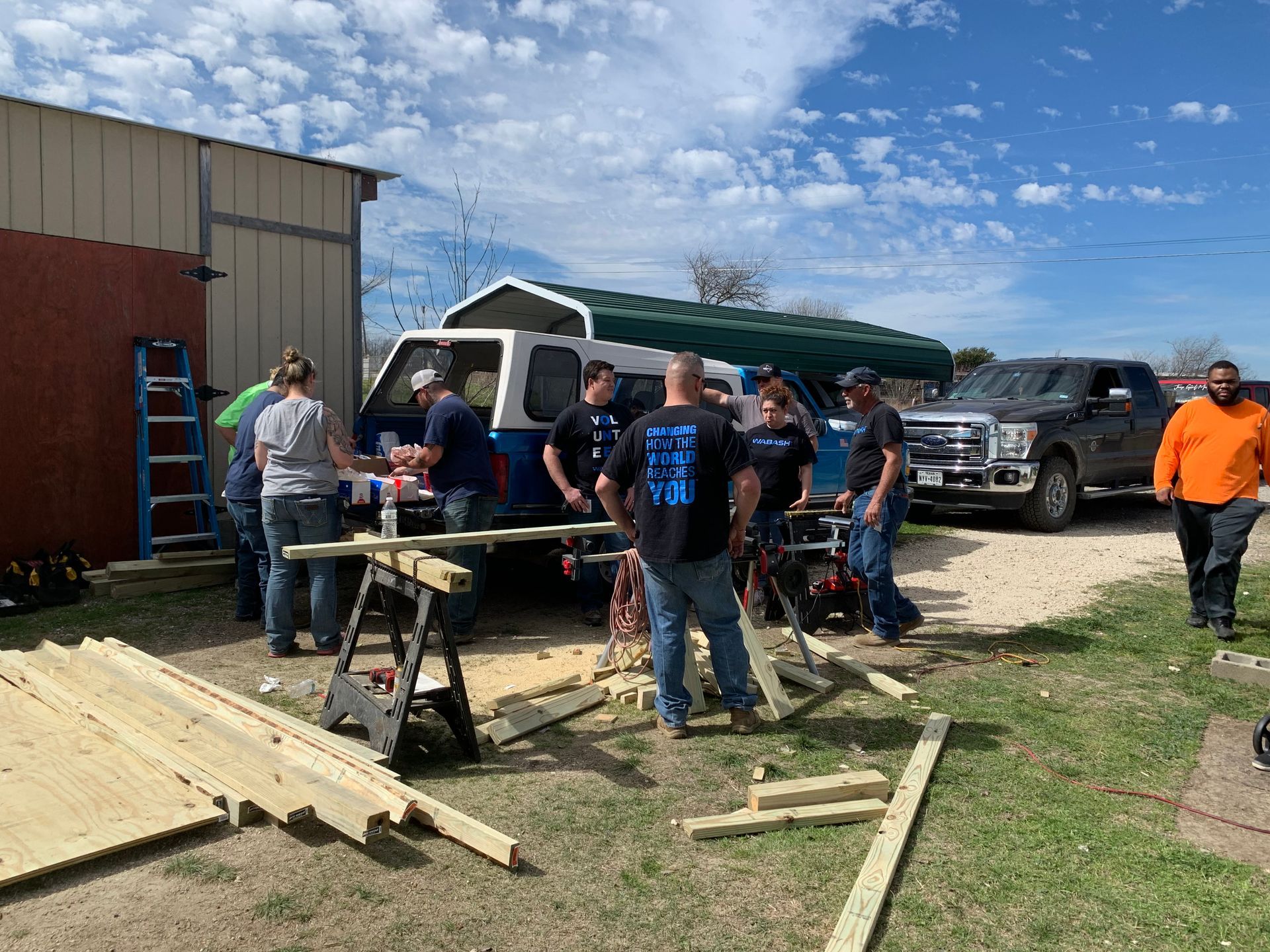 People gathered around a picnic table outside a barn, with trucks parked nearby and lumber on the ground.