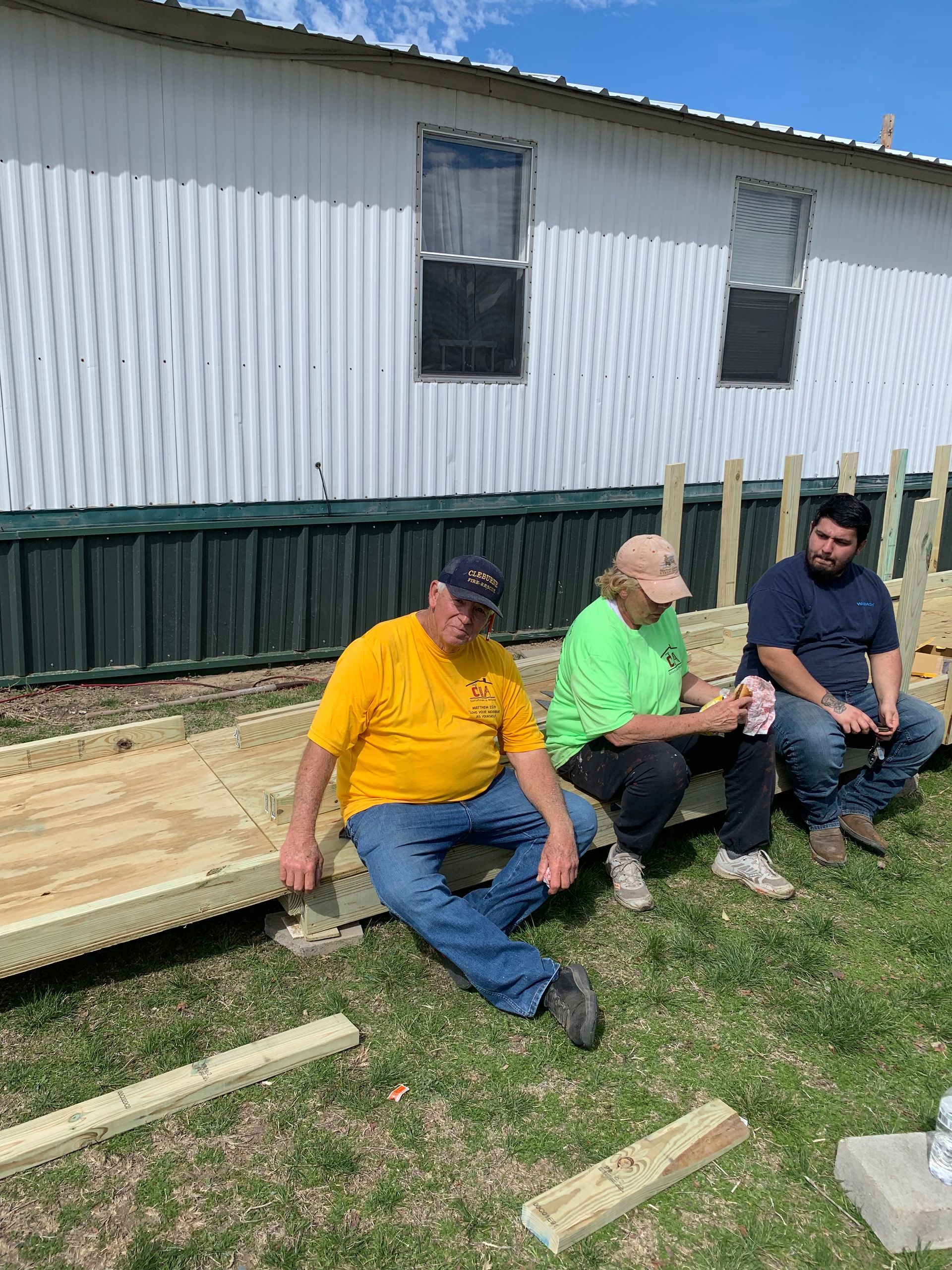 Three people sitting on a low wooden bench outside a white building with a green base, on a grassy yard.