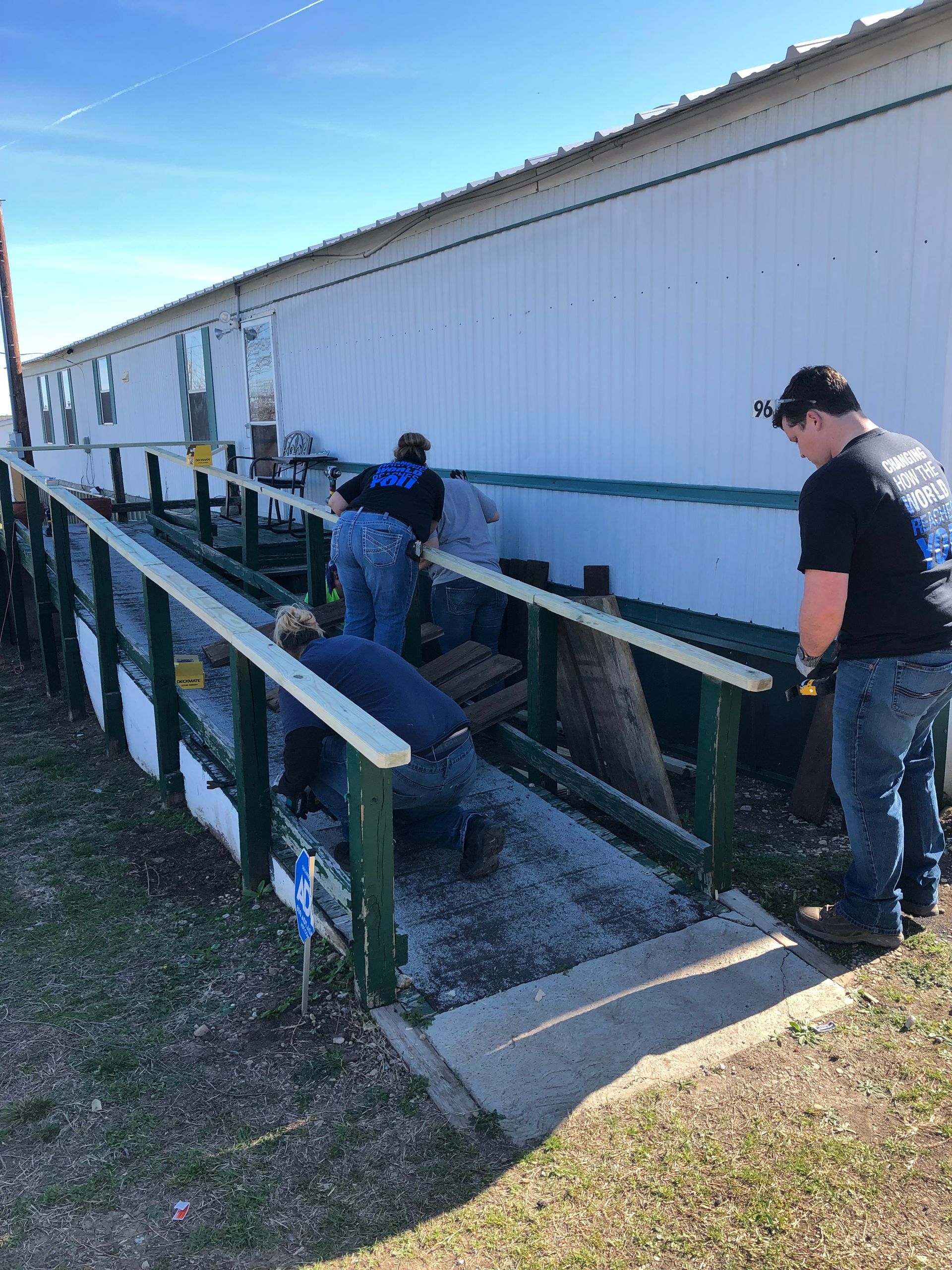 Workers on an outdoor ramp beside a white building, inspecting or repairing railings under bright sun