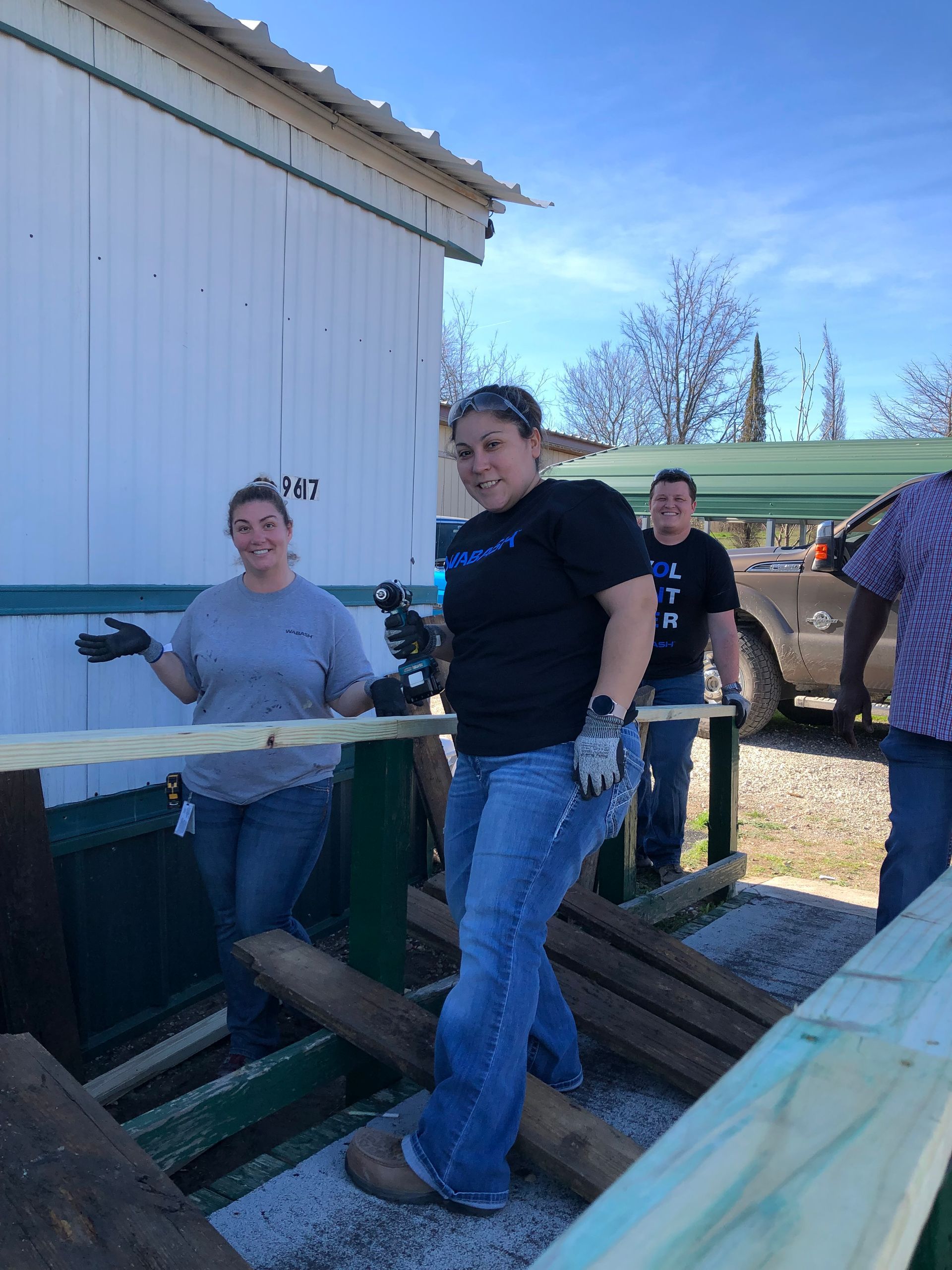 Three people standing on a farm trailer beside a white building, with one holding a tool.
