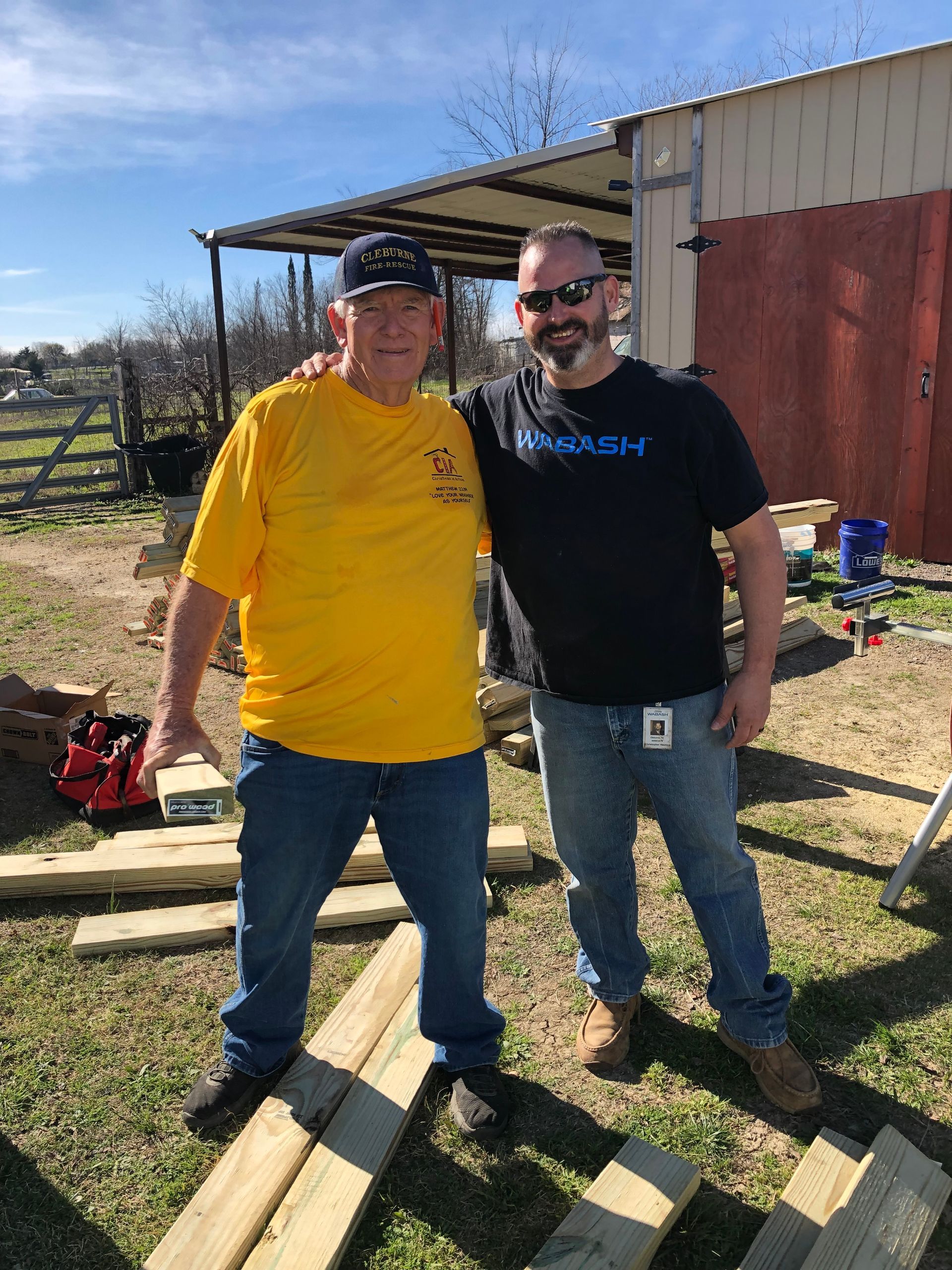 Two men standing outdoors by a barn and lumber piles, smiling in bright sunlight.