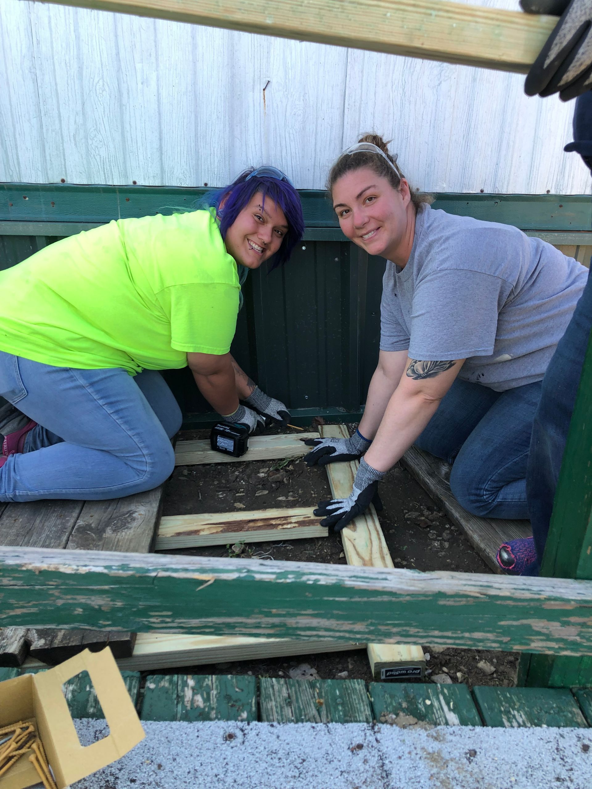 Two people kneel beside an open floor hatch, inspecting tools and wood framing under a structure.