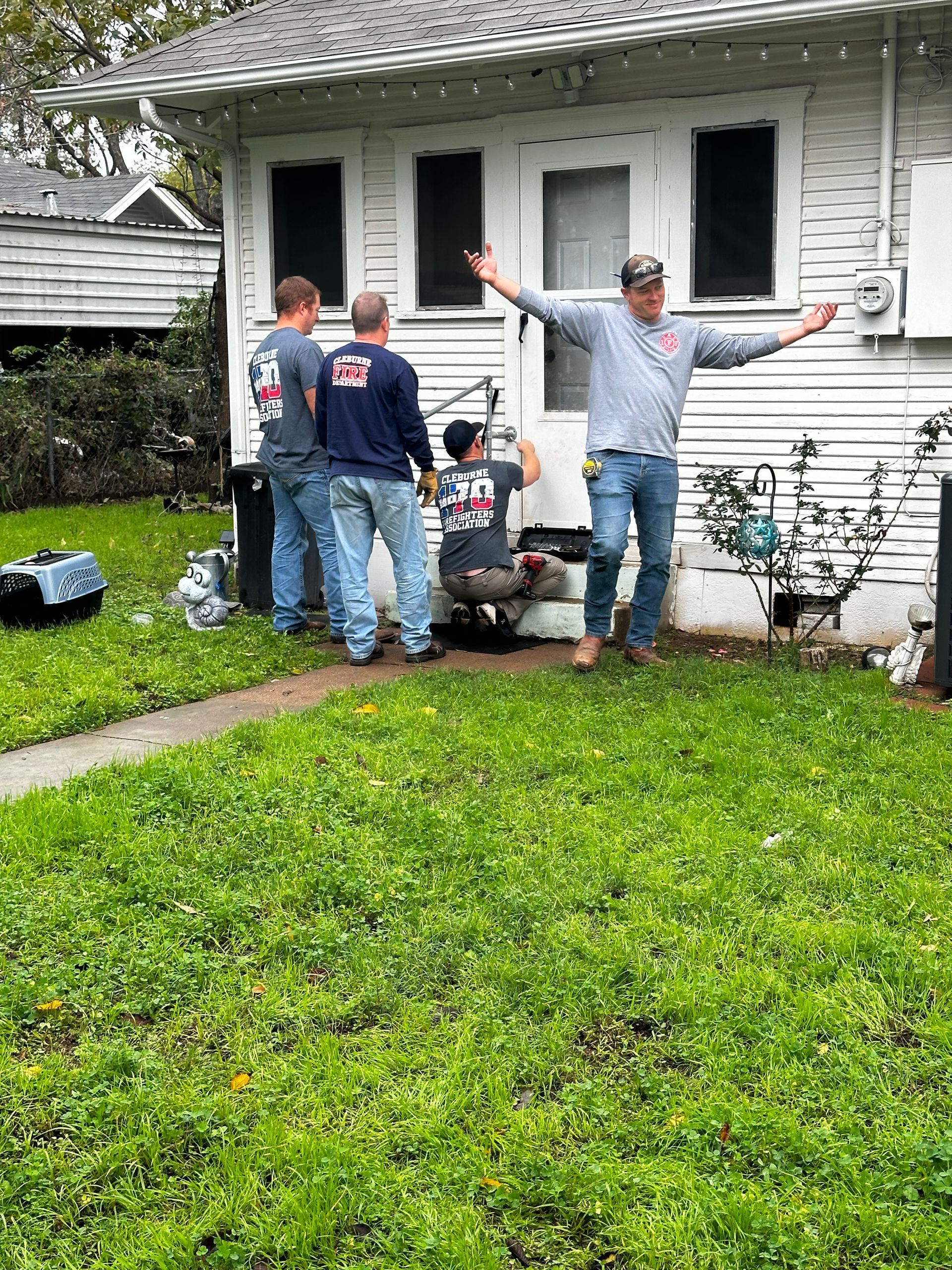 People standing outside a white house, one with arms raised, near a porch and lawn.
