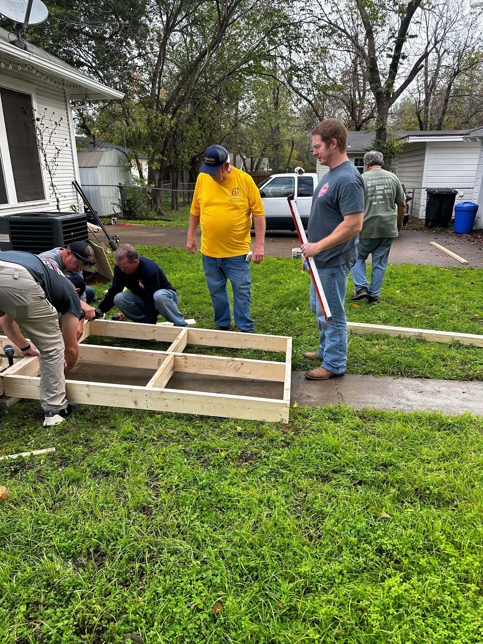 People assembling a wooden planter box in a front yard beside a sidewalk and house
