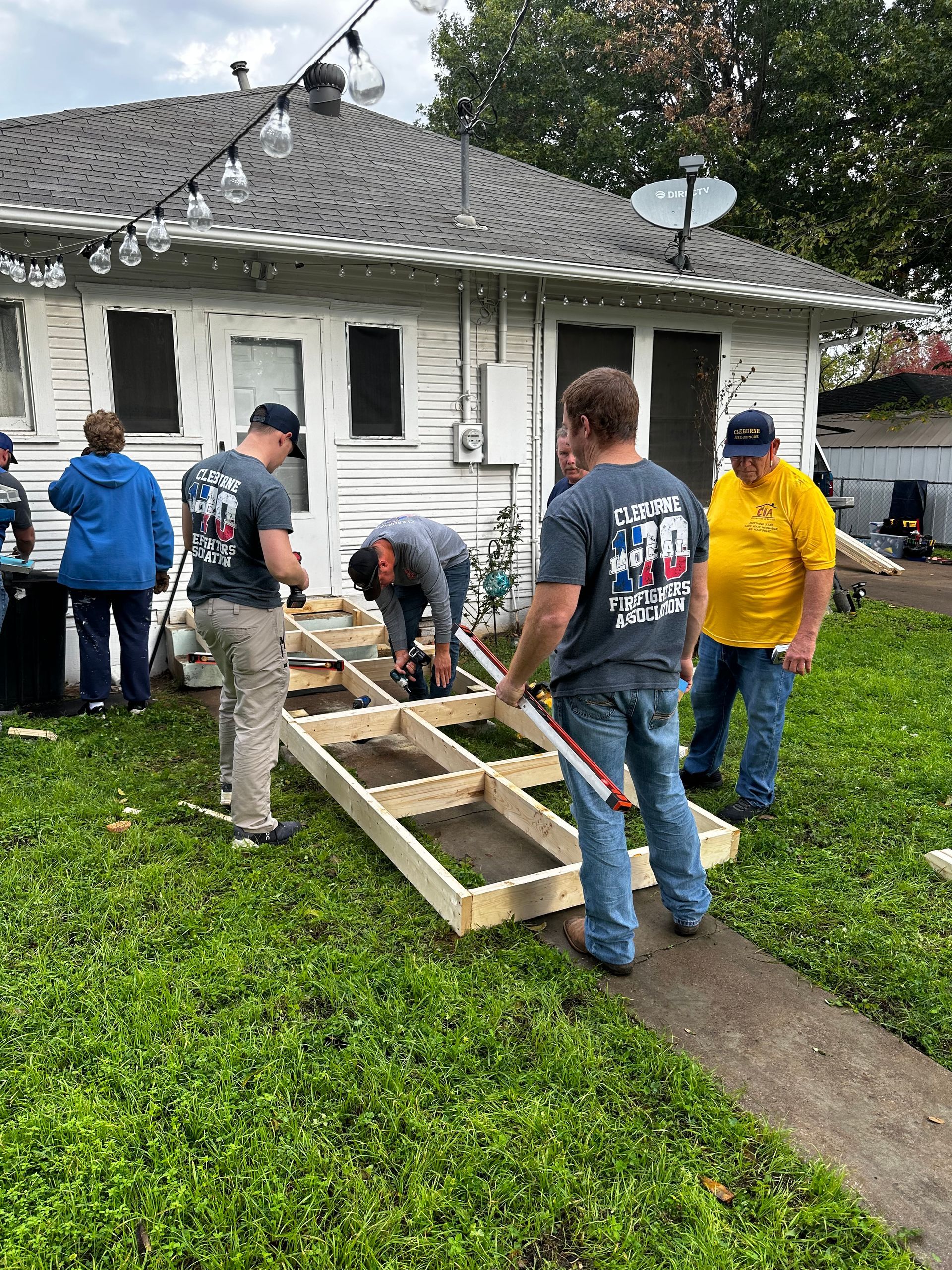 Volunteers carrying a wooden ramp frame toward a white house in a yard after rain
