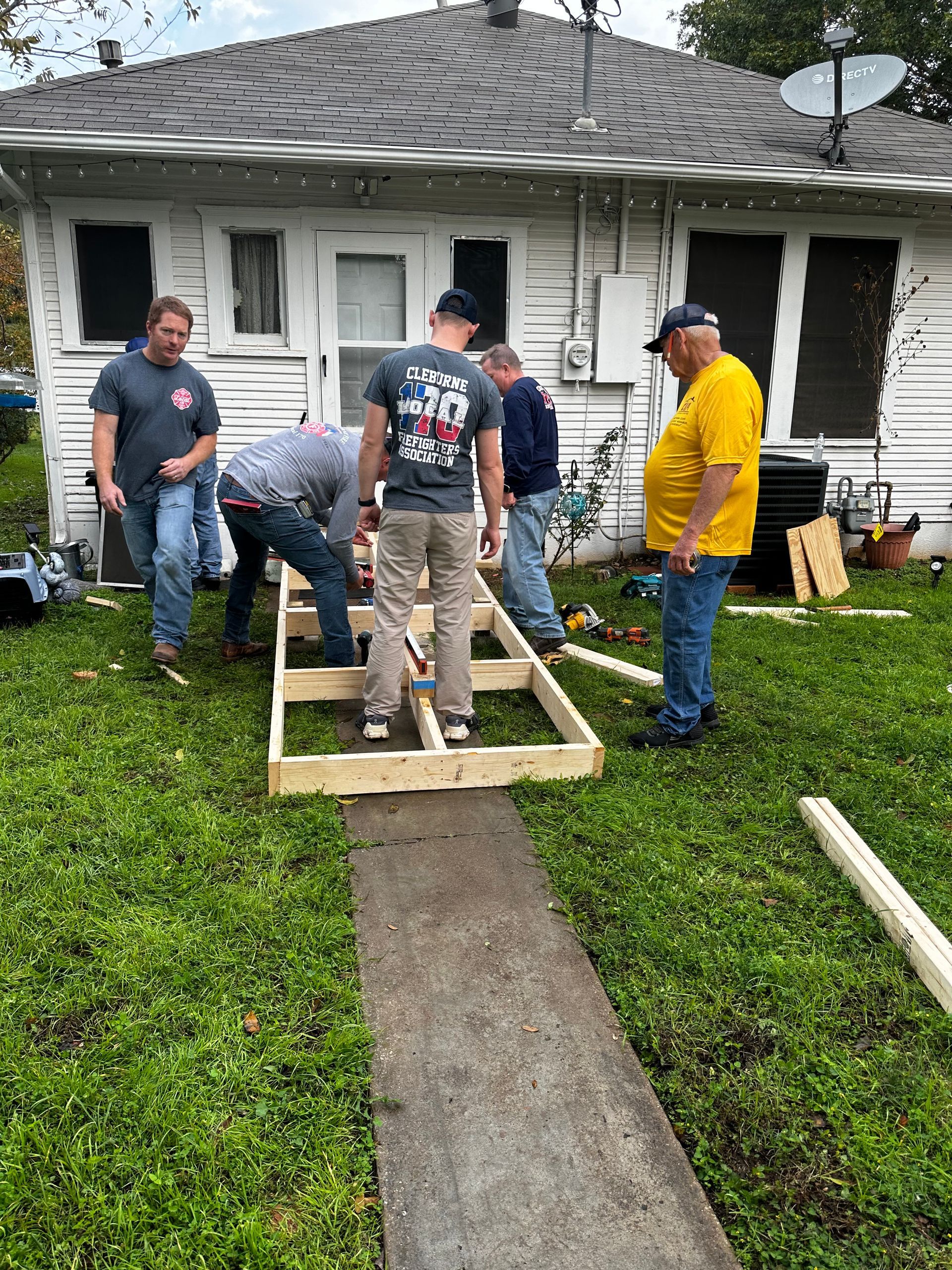 Five people building a wooden ramp outside a damaged white house, with tools and a concrete path in front
